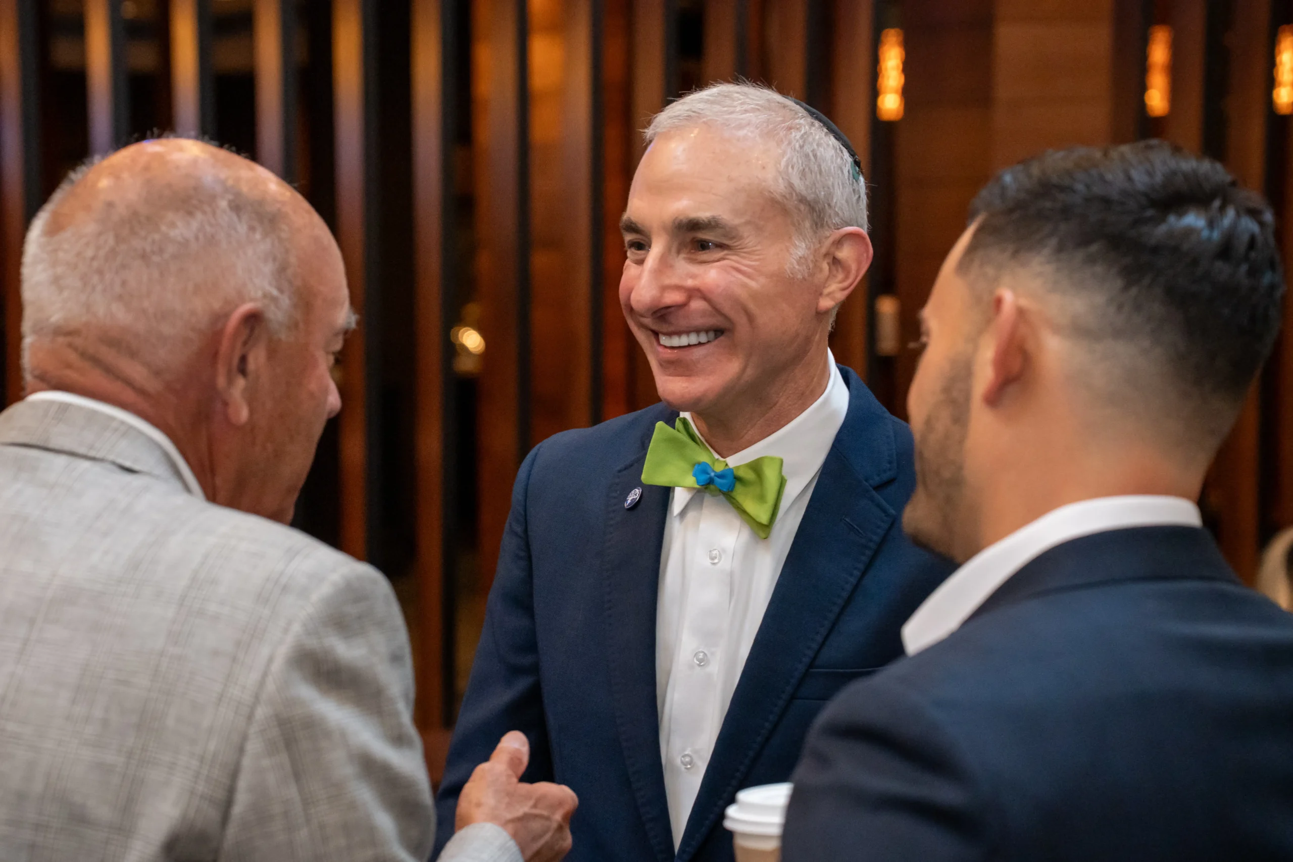Three people in formal attire converse indoors. One person wears a green bow tie. Wooden background elements are visible. Smiling and engaging moment captured.