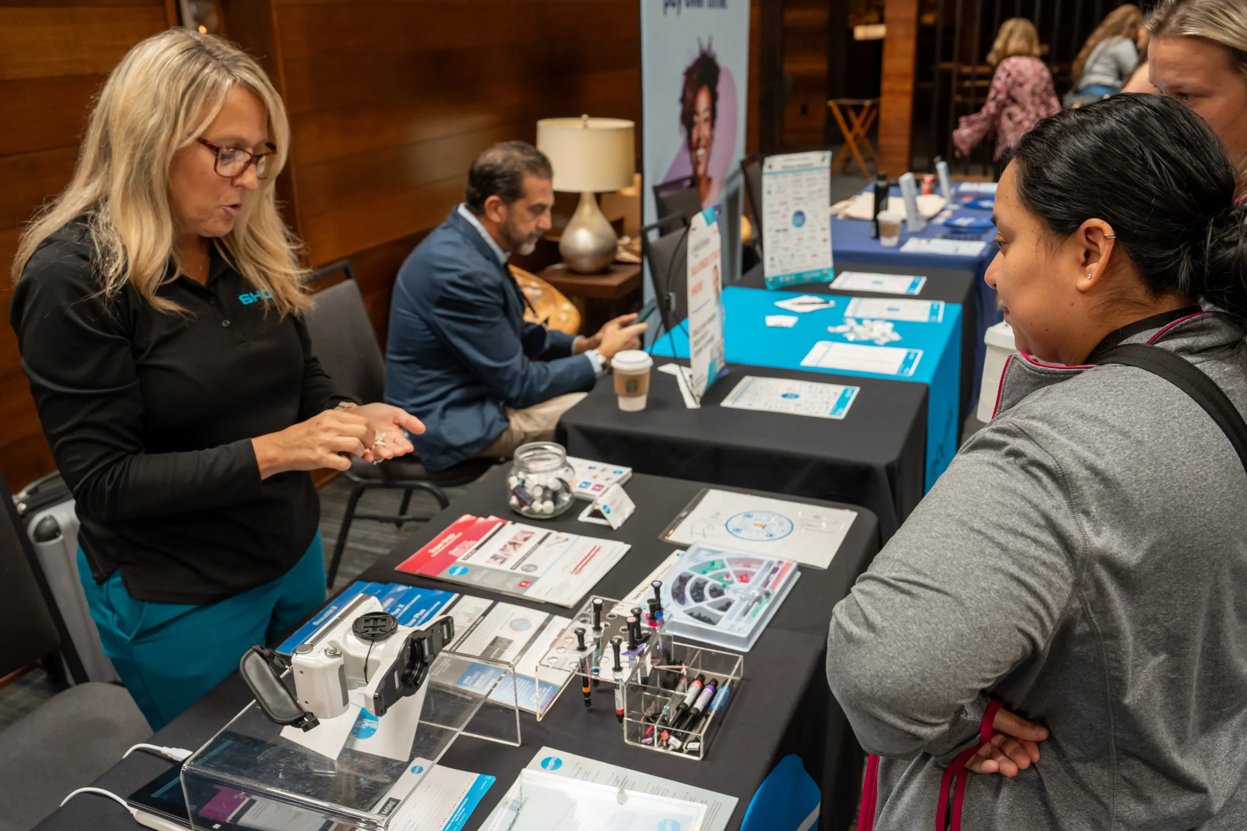Two people at a technology exhibit table, discussing gadgets and brochures. A person in the background is seated, using a tablet.