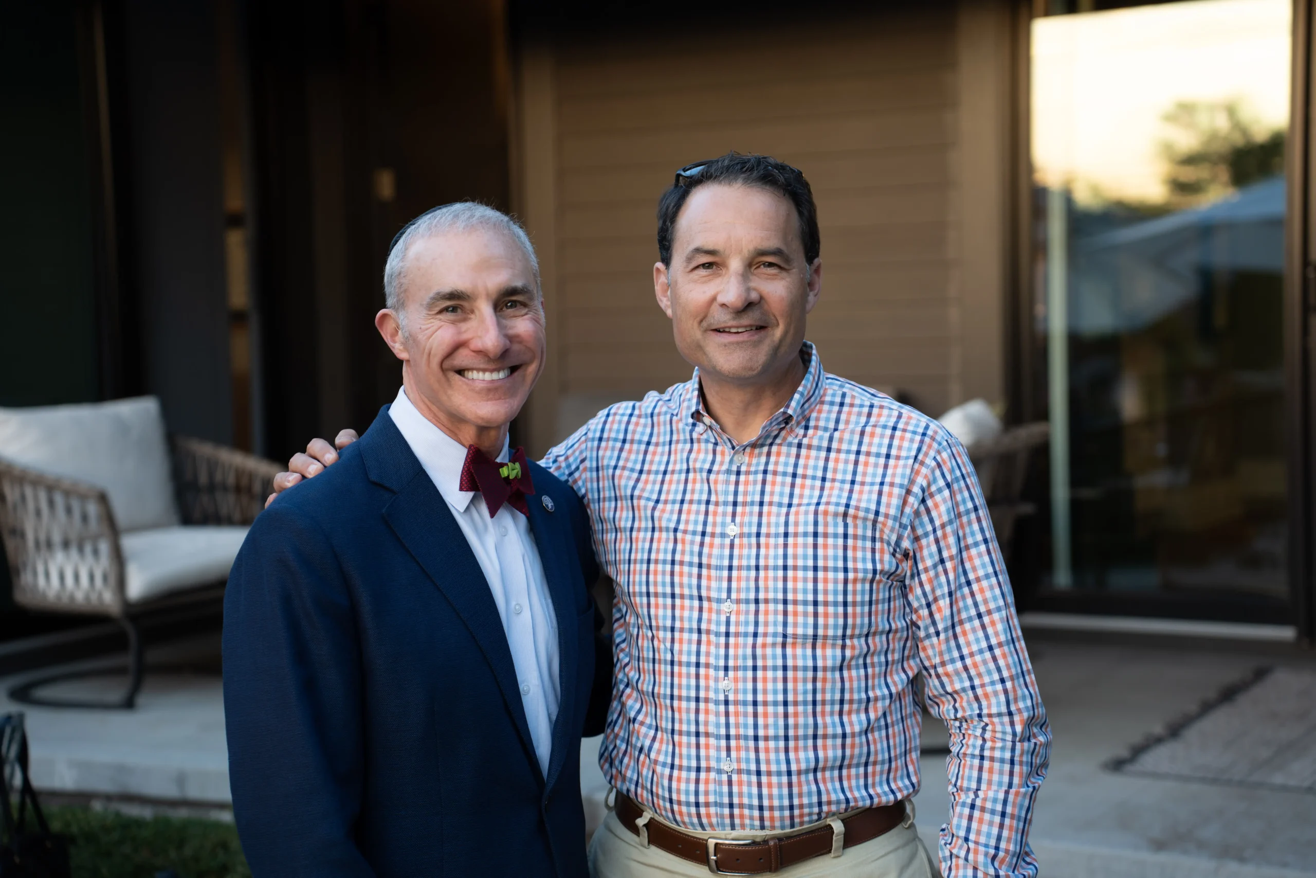Two smiling people stand close together outdoors, one in a suit and the other in a checkered shirt. Patio furniture visible in background.