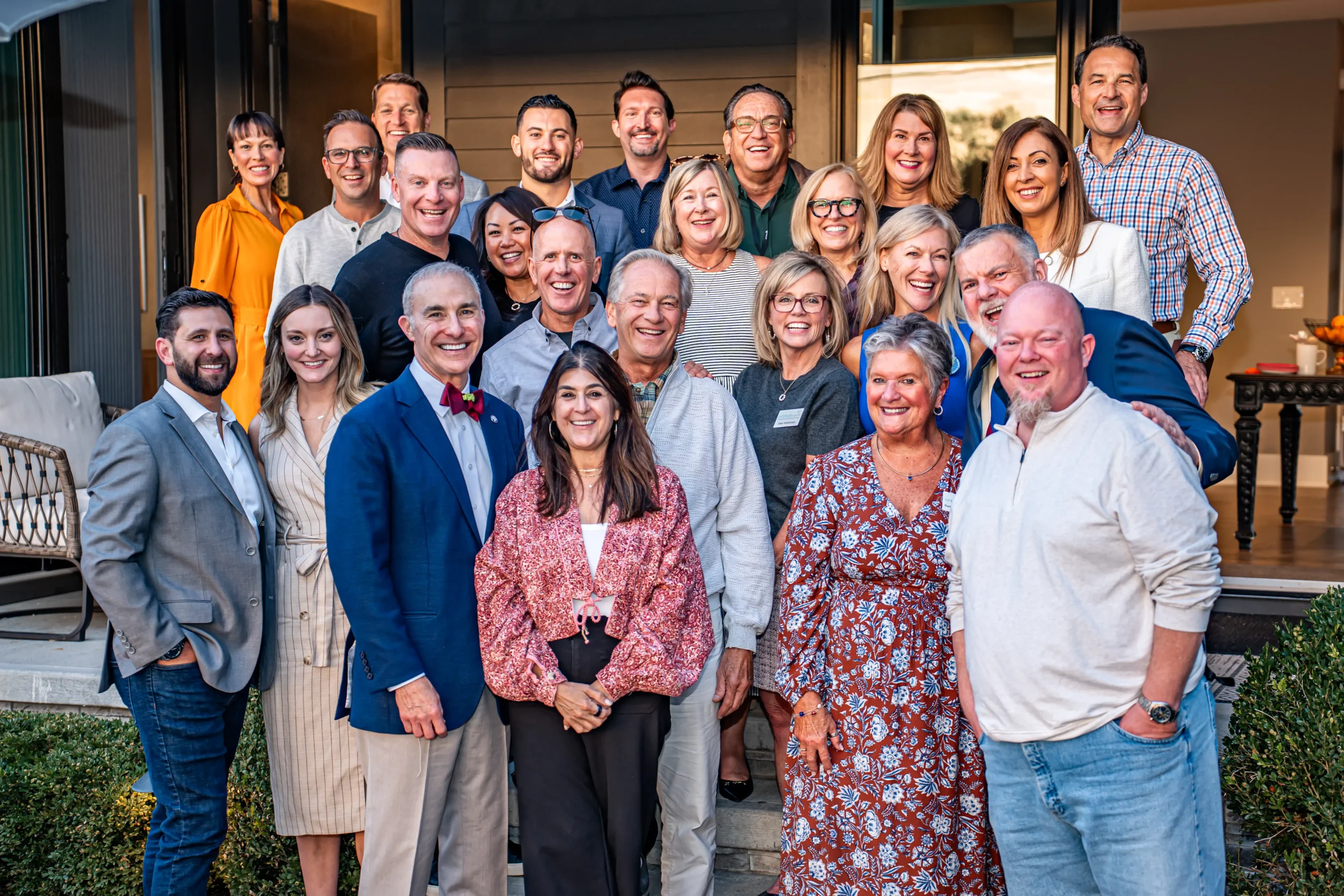 A group of people smiling and posing together outdoors, in front of a modern building, under a clear sky.