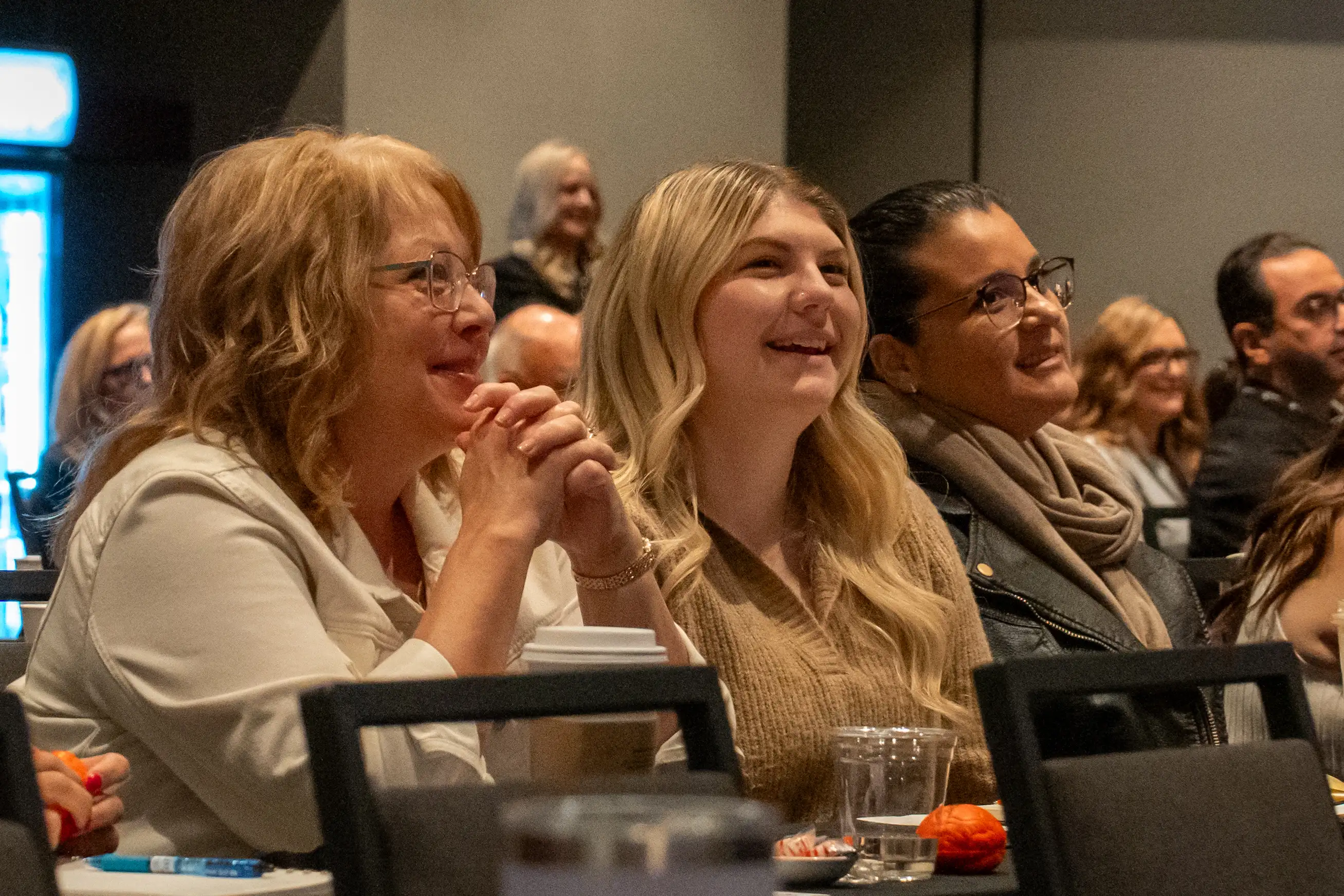 Three people sitting closely in an audience, smiling attentively during an indoor event. They appear engaged and are holding drinks and notepads.