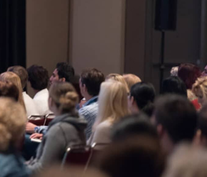 A group of people seated in a room, attentively listening to a presentation or speaker, suggesting a seminar or conference setting.
