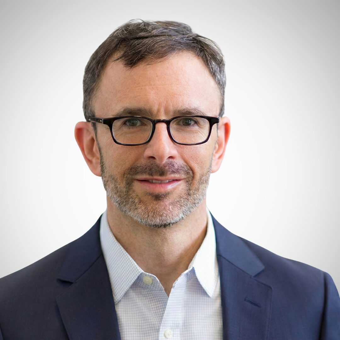 A person with glasses wearing a suit and shirt, smiling against a plain background. No identifiable landmarks or historical buildings are visible.