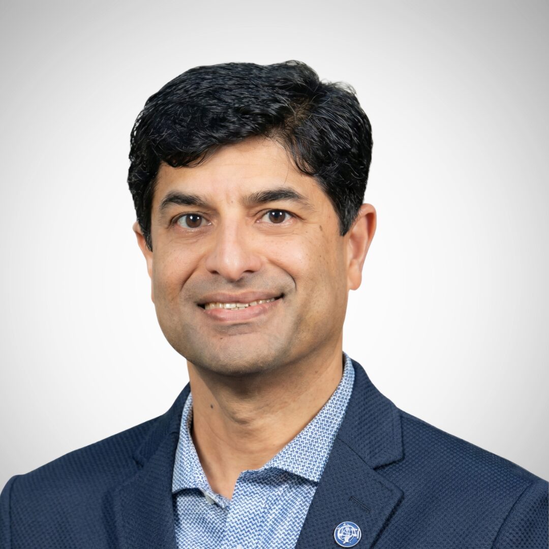 A person in a blue blazer and patterned shirt smiles against a plain white background.