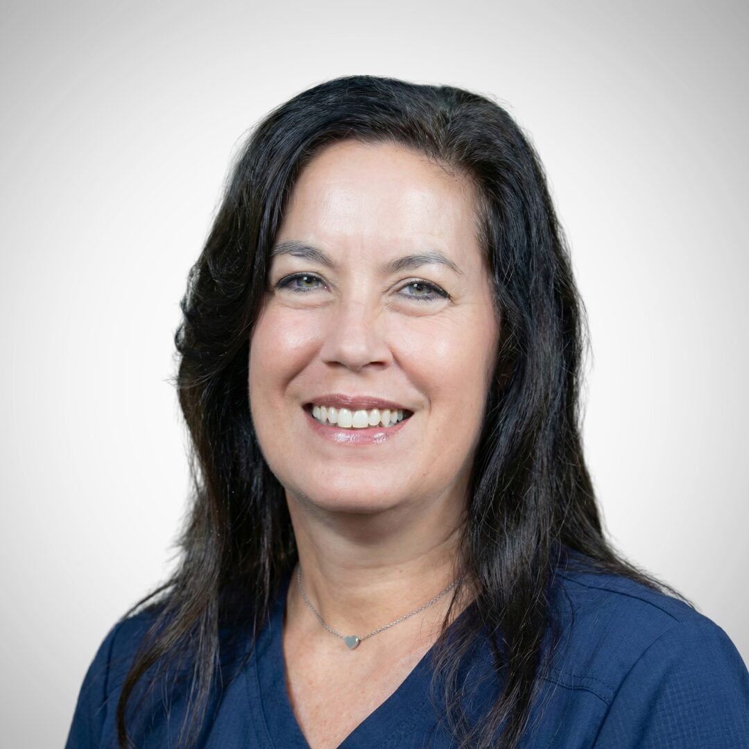 A person with long dark hair and a friendly smile is wearing a dark blue top. The background is plain and light-colored.