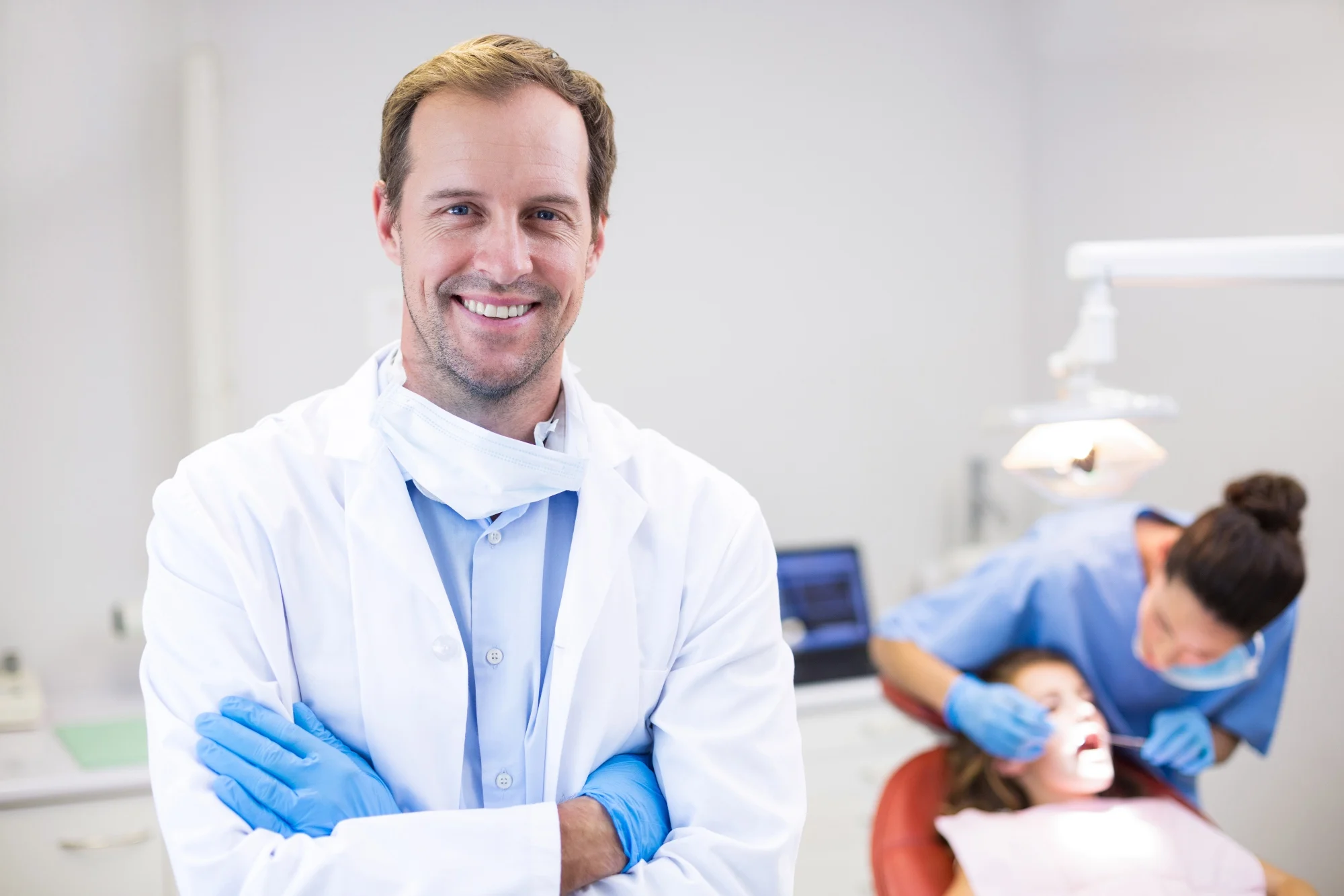 A person in a white coat smiles confidently, standing in a dental office, while another person provides dental care to a seated patient.