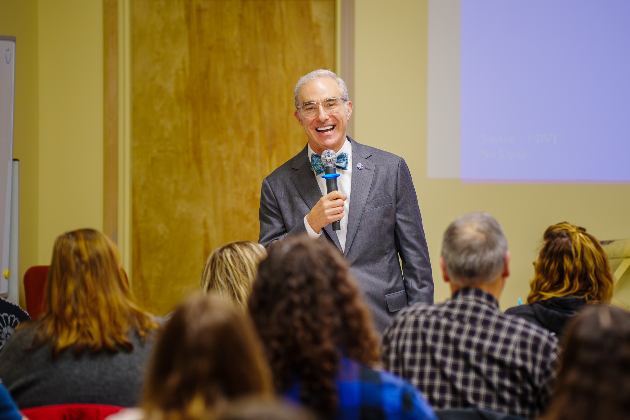 A person in a suit holds a microphone, speaking to a seated audience in a conference room setting.