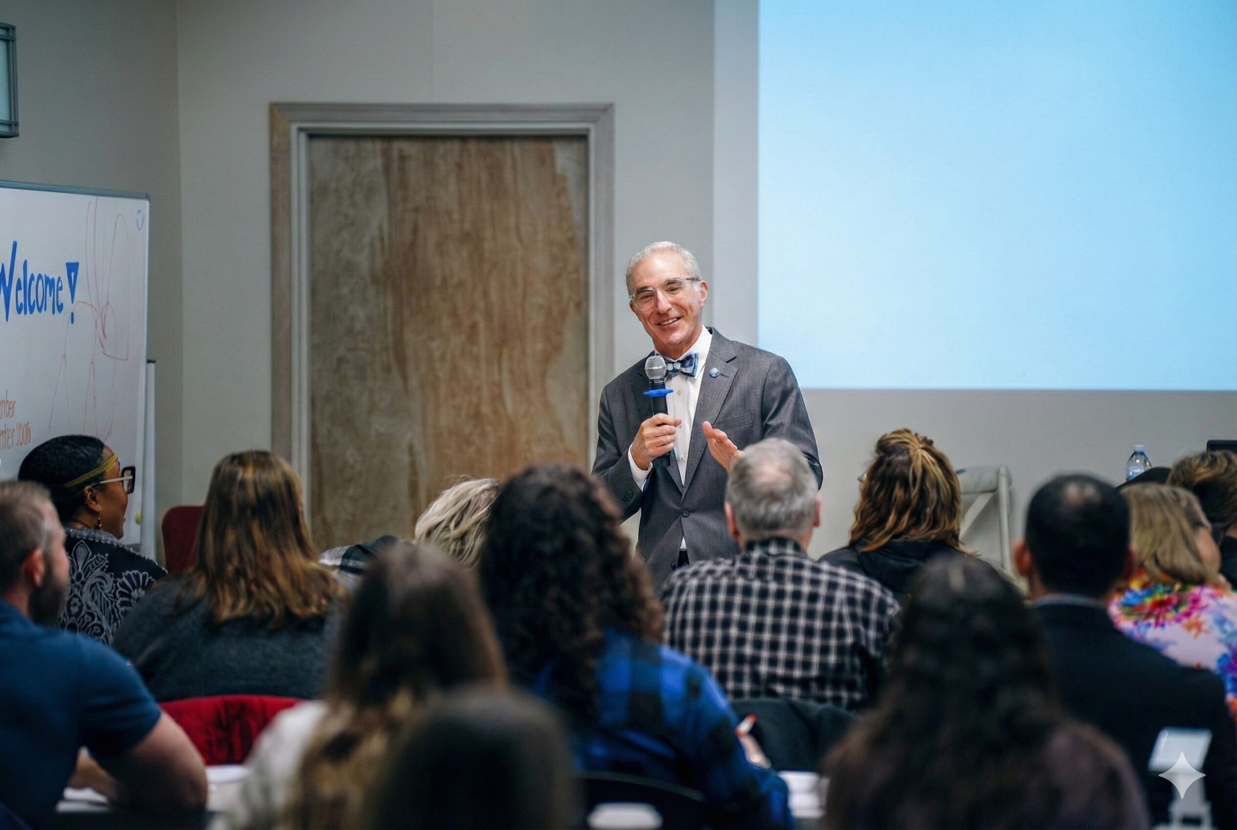 A person in a suit speaks to a seated audience in a conference room with "Welcome!" on a whiteboard.