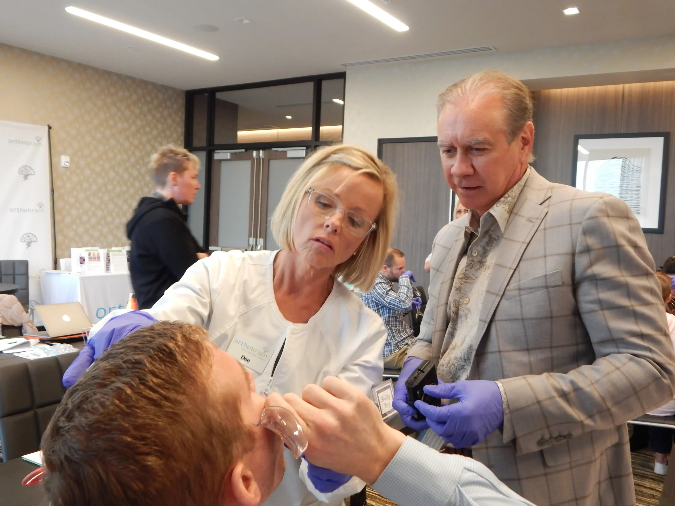 A person examines another's teeth while another watches in a modern conference room, with several people in the background.