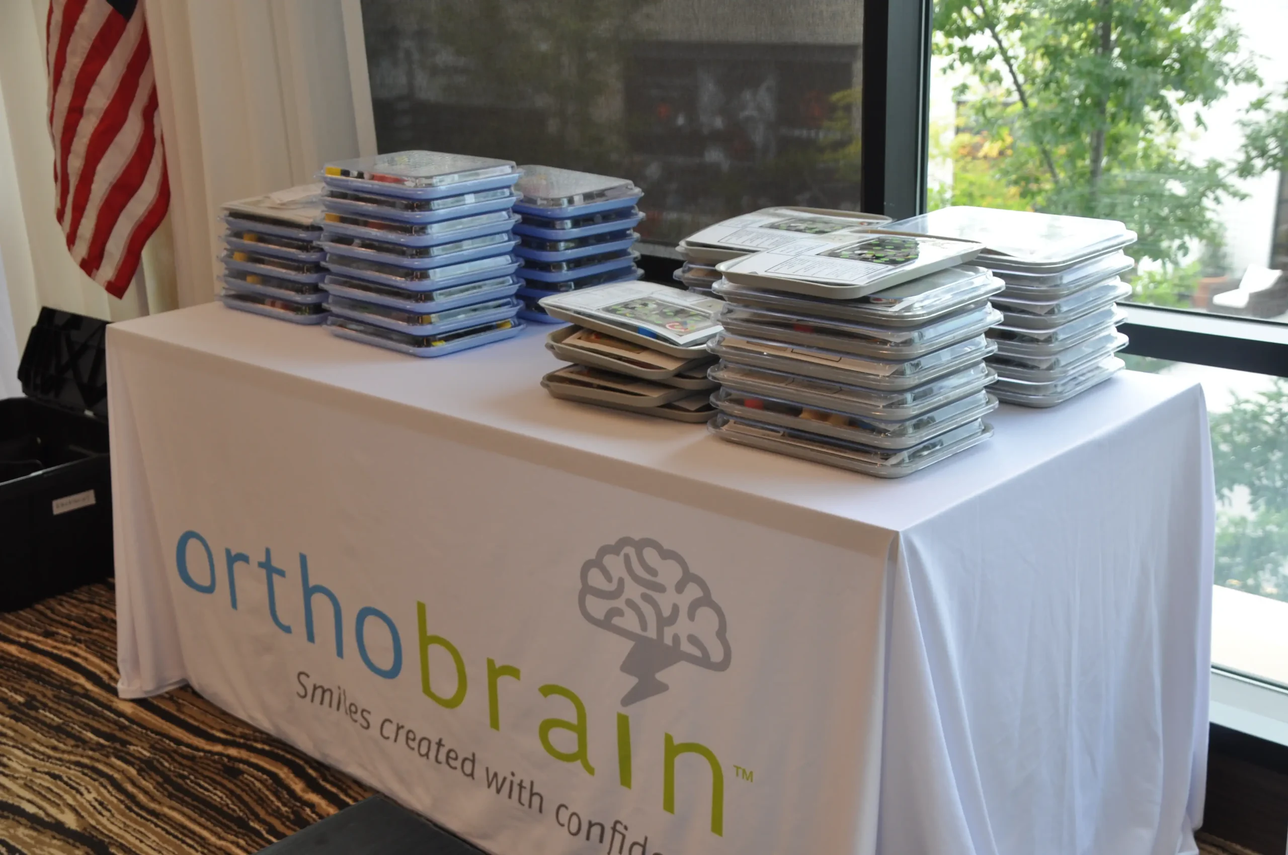 A table with "orthobrain" branding displays stacks of educational materials. An American flag stands nearby, with greenery visible through a window.