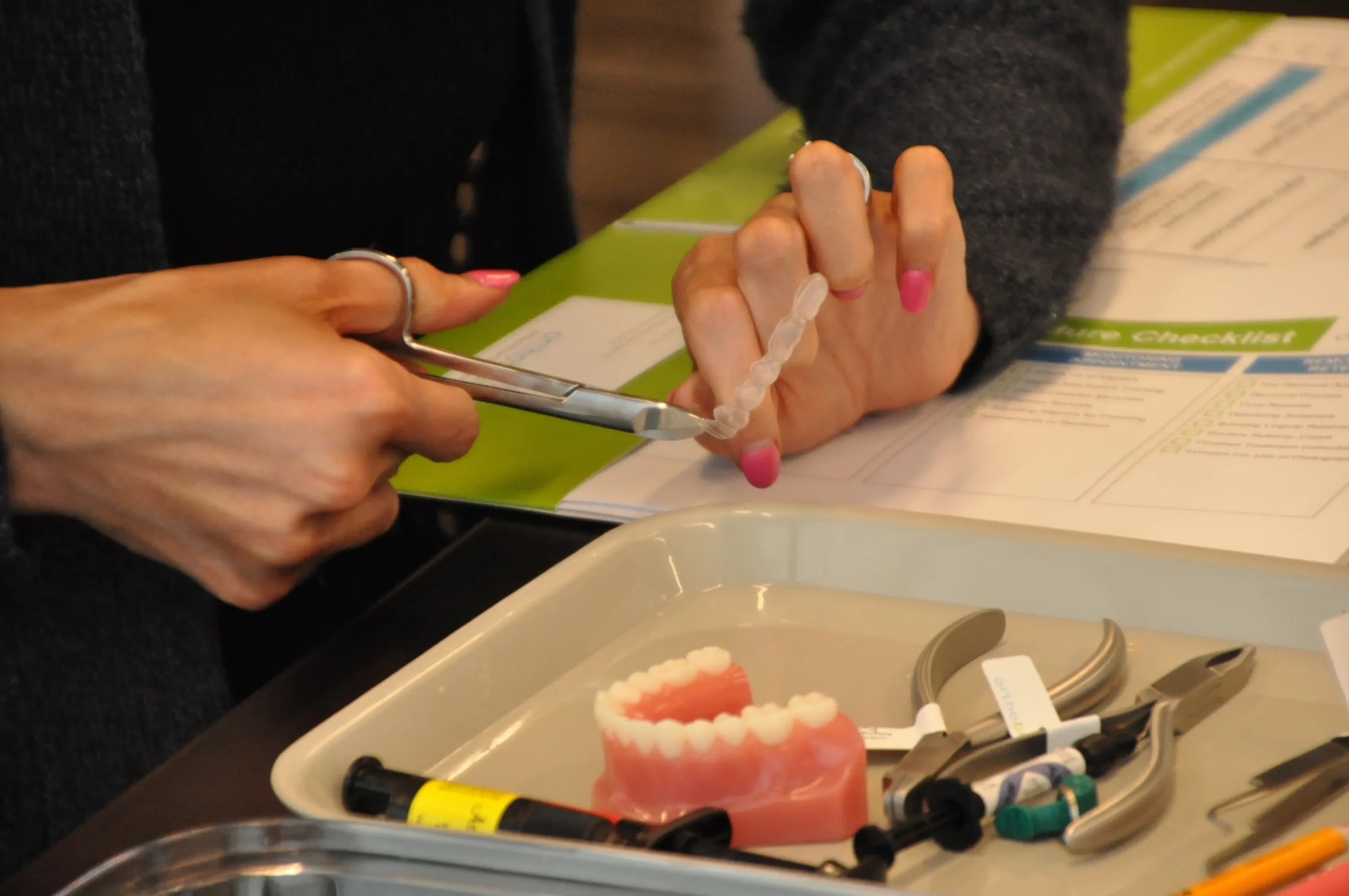 A person prepares a dental model with scissors, surrounded by dental tools and instructional papers in a clinical setting.