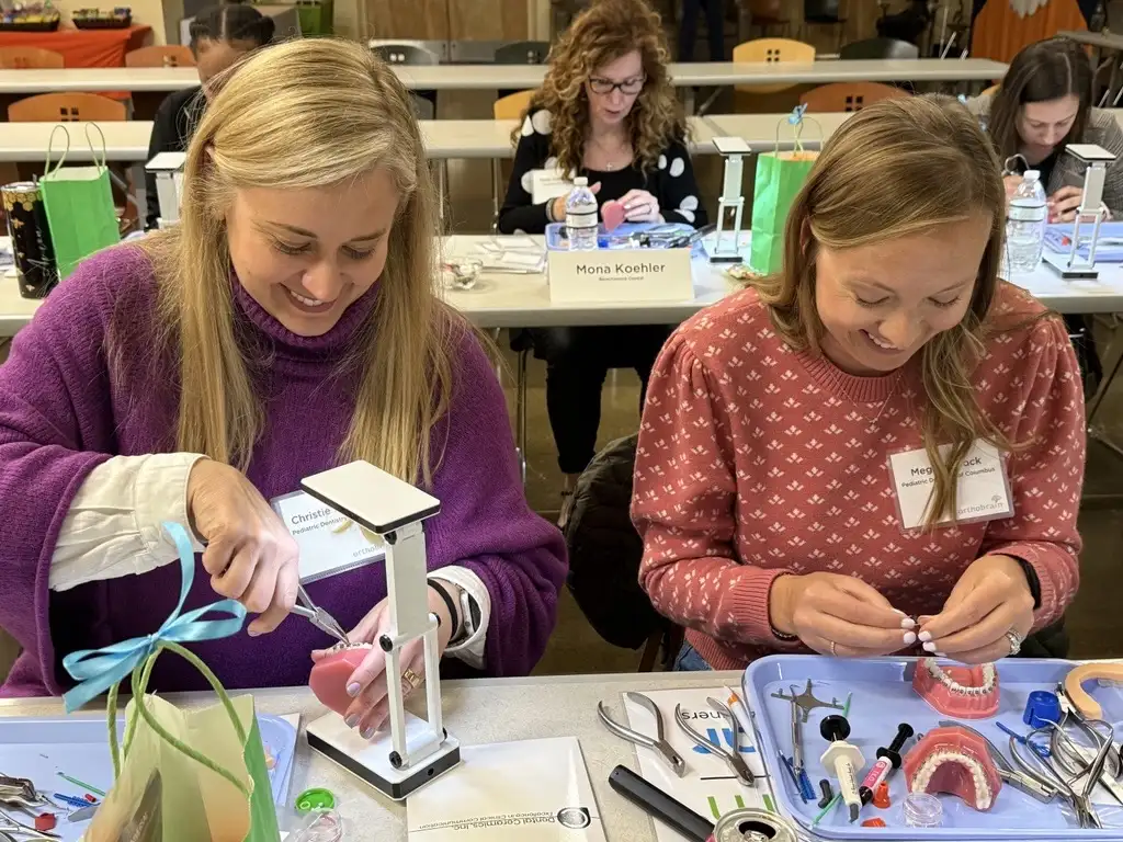 Two people work on dental models at a workshop, using various orthodontic tools. Others are engaged in similar activities in the background.