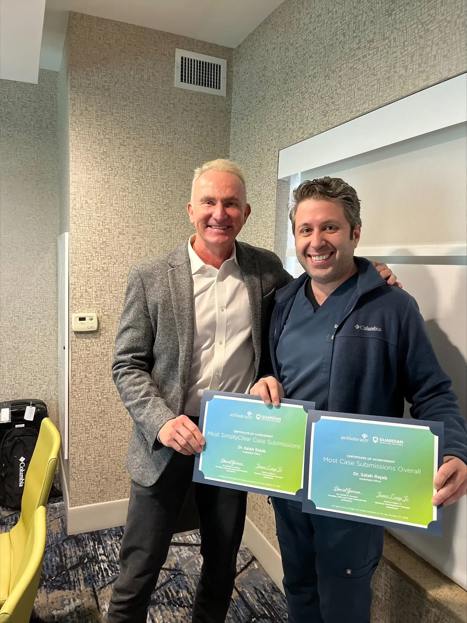Two people smiling indoors holding certificates, standing in front of a patterned wall. A bright yellow chair is visible on the side.