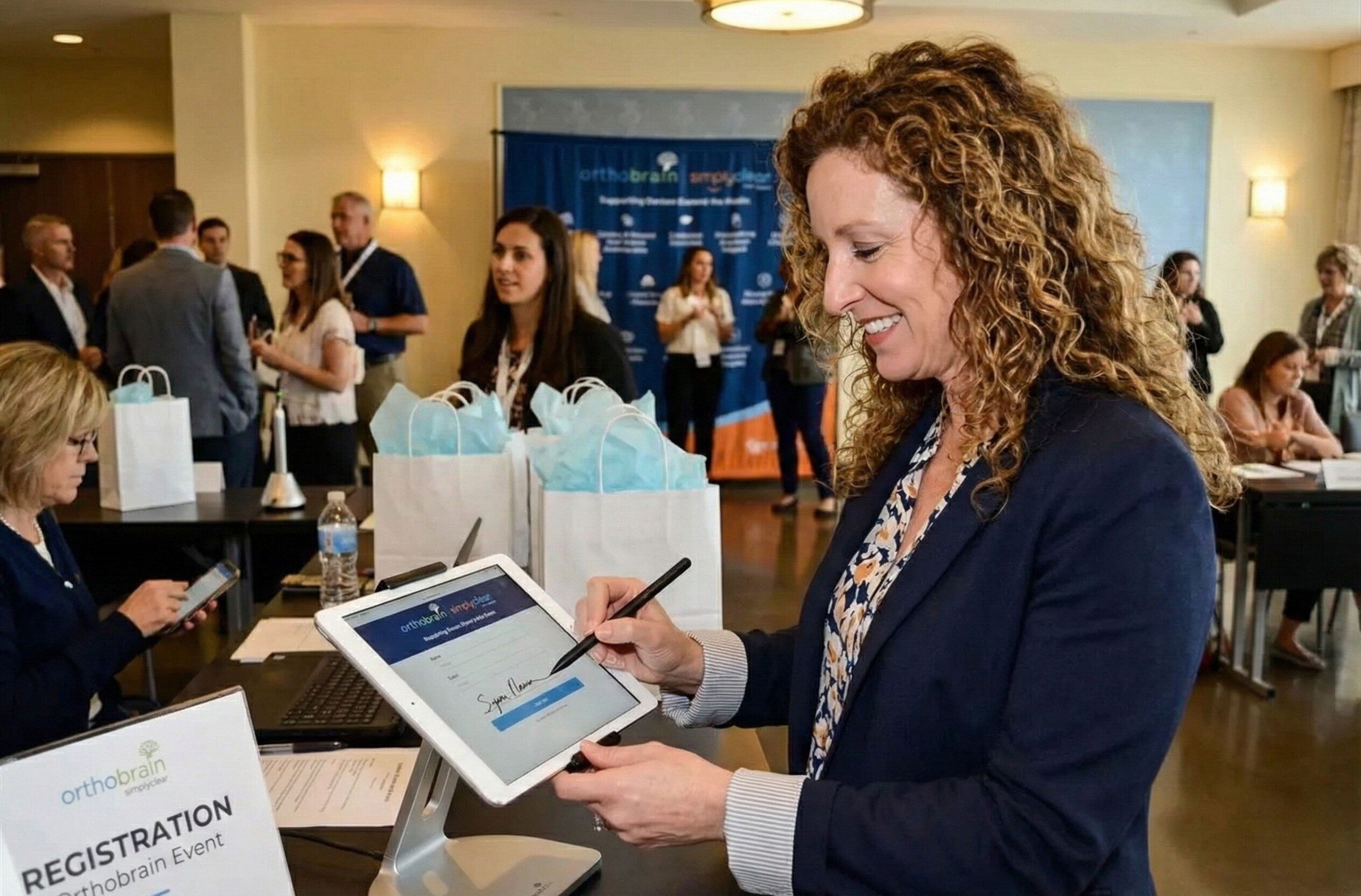 Person signing in at an event registration desk. Others gather and converse in the background. Bags are lined up on the table.