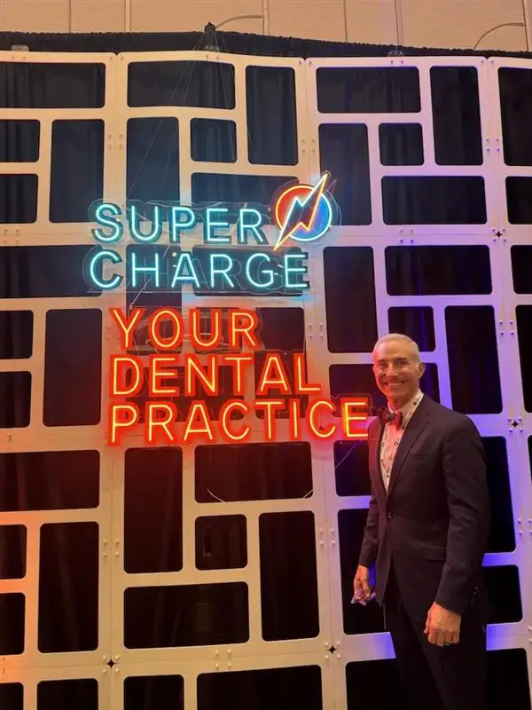 A smiling person stands beside a neon sign reading "Super Charge Your Dental Practice" on a decorative grid backdrop, suggesting a motivational event.